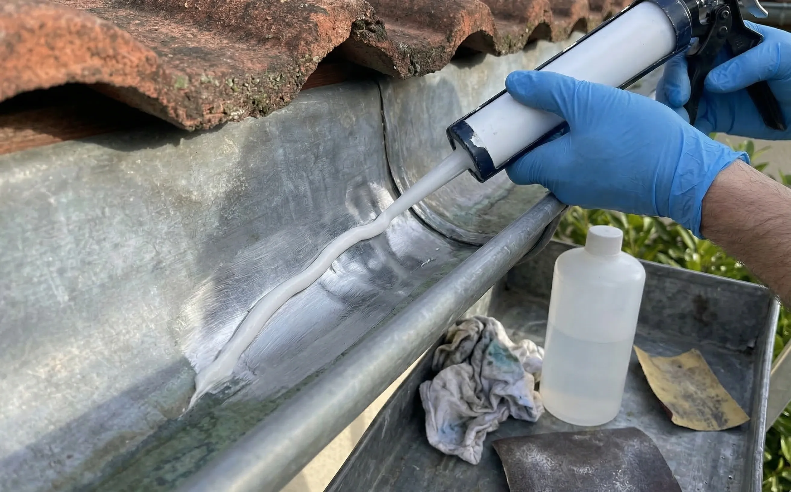 Mains gantées appliquant un mastic MS polymère au pistolet sur une fissure de gouttière zinc décapée