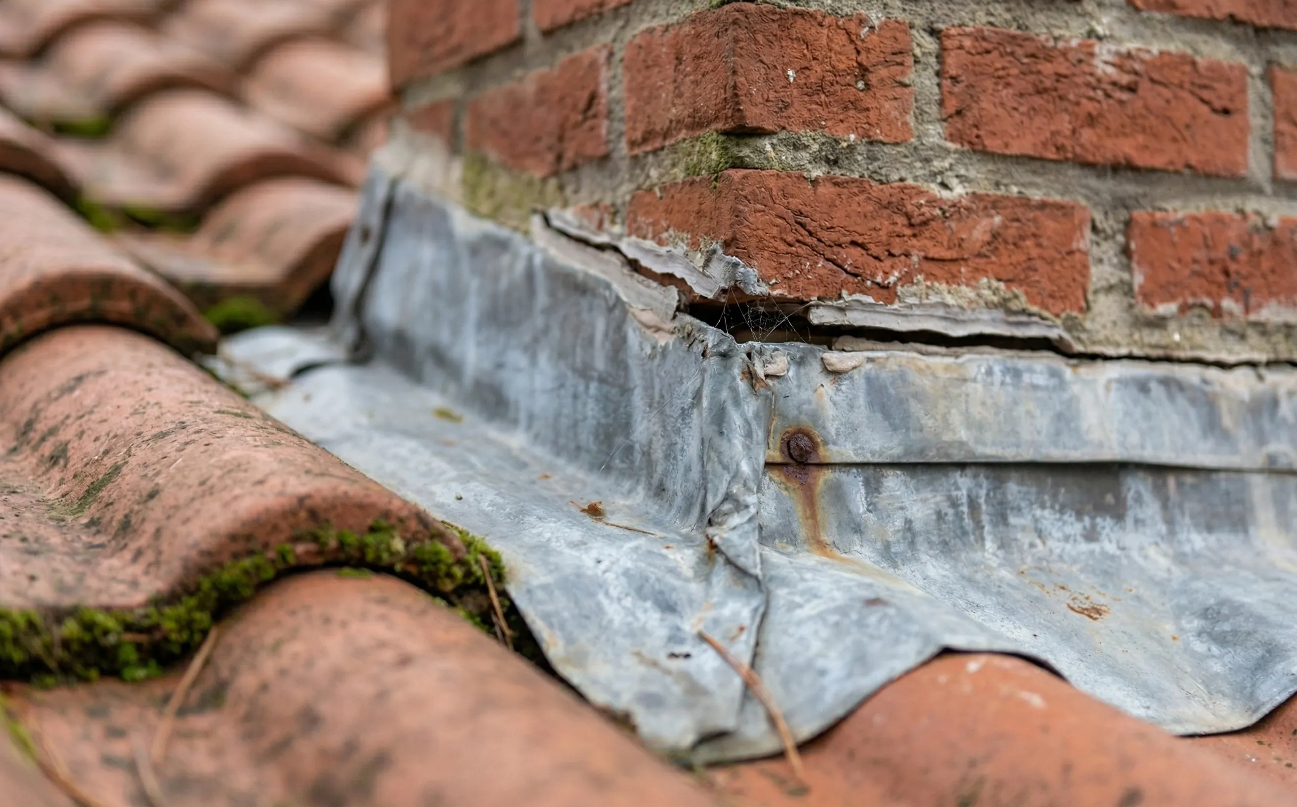Gros plan sur un solin de cheminée en zinc patiné, joint mastic partiellement décollé, tuiles mécaniques avec traces de mousse au raccord