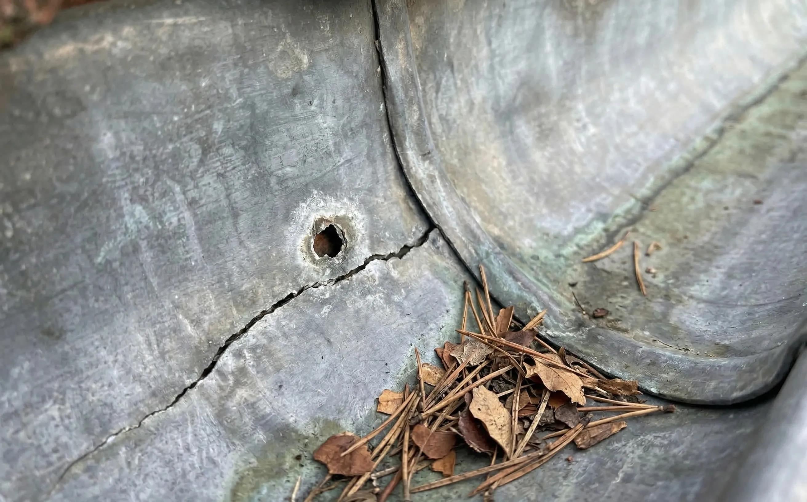 Trou de corrosion cerné de patine blanchâtre et fissure linéaire sur une gouttière zinc